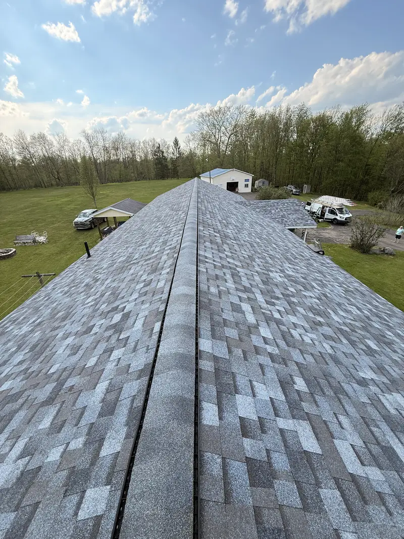 Architectural asphalt shingle roof photographed along the ridge, with open lawn, trees and an outbuilding beyond.