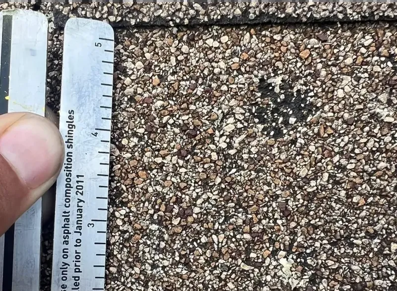 Close-up of a measuring gauge held against an asphalt shingle beside a small dark mark.