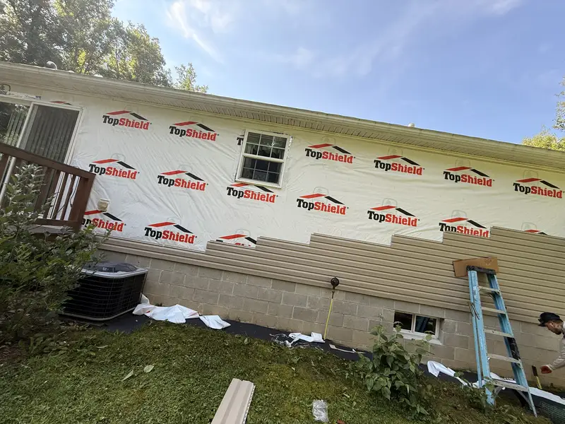 House wall wrapped in housewrap part-way through a siding installation, with lap siding run along the lower course, a worker beside a stepladder and offcuts on the ground.